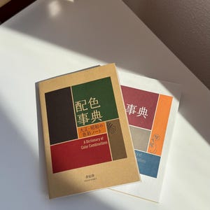 Könnte beinhalten: Zwei rechteckige Bücher mit beigen und weißen Einbänden, die geometrische Blöcke in Dunkelbraun, Grün, Rot und Orange aufweisen. Das vordere Buch zeigt den Text "配色事典", "大正・昭和の色彩ノート" und "A Dictionary of Color Combinations" in Weiß und Schwarz.