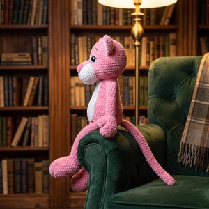 Peut inclure: Une peluche crochetée rose en forme de panthère avec un museau et un ventre blancs, de grands yeux sombres et une longue queue. L'animal en peluche est fabriqué à partir de fil texturé et est positionné en position assise avec ses membres étendus.