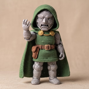 Może przedstawiać: Szydełkowa figurka amigurumi przedstawiająca postać w zielonej pelerynie z kapturem, zielonej tunice i srebrnej zbroi. Figurka posiada maskę o metalicznym wyglądzie, brązowy pasek z małą torebką oraz złote, okrągłe zapięcia przy pelerynie.