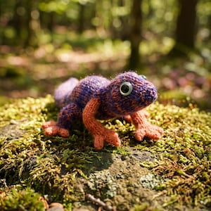 May include: A knitted lizard figurine featuring a textured purple and orange body with prominent plastic eyes. The creature has four distinct limbs with splayed toes and a long tail, crafted from yarn with visible stitching throughout the surface.