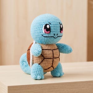 Op de afbeelding: Een lichtblauw gehaakt knuffeltje in de vorm van een schildpadachtig wezen met een beige schild op de rug. Het figuurtje heeft grote zwart-roze ogen, een klein mondje en een gekrulde staart. De textuur van het garen is overal goed zichtbaar.