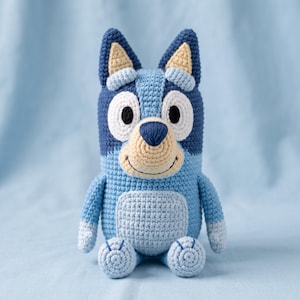 Op de afbeelding: Een handgehaakt knuffeldier van een blauwe hond. Het figuur heeft donkerblauwe oren en een donkerblauwe bovenkant van het hoofd, een lichtblauw lichaam en een crèmekleurige snuit en binnenkant van de oren. Het heeft grote witte oogvlekken.