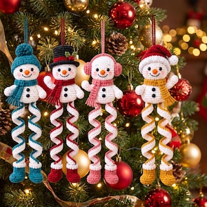 May include: Four crocheted snowman ornaments with dangling spiral legs. Each snowman features a unique hat and scarf set in teal, red, pink, or mustard yellow, with black button eyes, a small orange nose, and two black buttons on the torso.