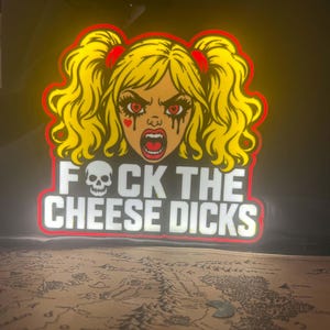 May include: A glowing LED sign featuring a cartoon character with yellow pigtails, heart-shaped eyes, and black makeup streaks. Below the character, the text reads FOCK THE CHEESE DICKS, with a white skull replacing the letter O in FOCK.