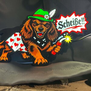 May include: A light-up sign featuring a brown dog wearing a green hat with a white feather and a black bandana with white bones. The dog wears white shorts with red hearts, holds a lit stick of dynamite, and has a speech bubble saying Scheiße!!