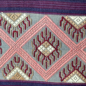 Può includere: Un tessuto intrecciato con un motivo geometrico ripetitivo di grandi rombi rosa su sfondo grigio chiaro. Il design include un intricato ricamo in filo rosso, beige e bianco, con strisce orizzontali viola scuro nella parte superiore e inferiore.