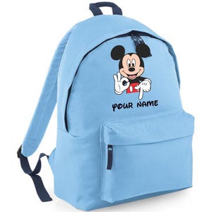 May include: A light blue backpack featuring a graphic of Mickey Mouse making an okay hand gesture and pointing downwards. Below the character, the text "YOUR NAME" is printed in black. The bag has dark blue zips, straps, and a top handle.