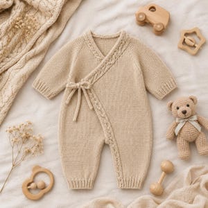 Op de afbeelding: Een beige gebreide baby-romper met een overslag aan de voorzijde, voorzien van een kabelgebreide rand langs de opening en een striksluiting aan de zijkant. Het kledingstuk heeft lange mouwen met geribbelde boorden en geribbelde broekspijpen.