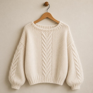 Peut inclure: Un pull à manches longues de couleur crème avec une texture en tricot épais, une large encolure ronde et un motif de torsades vertical descendant au centre de la poitrine et le long de chaque manche, suspendu sur un cintre en bois.