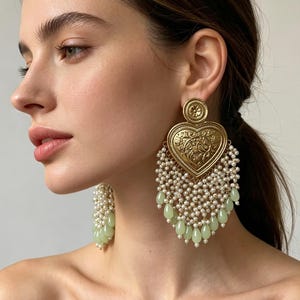 May include: A pair of gold-toned heart-shaped earrings featuring an intricate embossed floral pattern. The earrings are adorned with multiple strands of small white seed pearls and finished with dangling pale green teardrop-shaped beads.