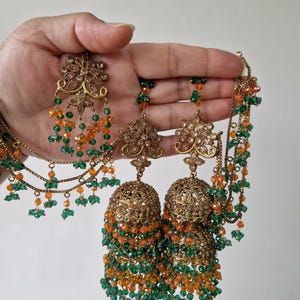 May include: A gold-toned jewellery set featuring a pair of ornate jhumka earrings and a matching multi-strand necklace. The pieces are decorated with intricate metalwork and dangling orange and green faceted beads in a tiered arrangement.