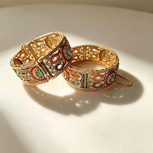 May include: Two gold-toned hinged bangles featuring intricate openwork metal patterns. The bracelets are decorated with small, colourful inlaid stones in shades of red, orange, green, and light blue, accented with tiny white beads along the edges.