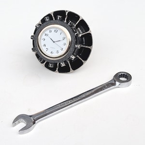 May include: A round clock with a white face and black numbers, set in a black and silver decorative frame. A silver wrench is positioned below the clock on a white surface.