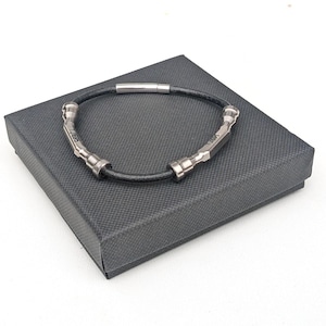 Könnte beinhalten: Ein schwarzes Lederarmband mit silbernen Akzenten. Das Armband wird in einer schwarzen Box präsentiert.