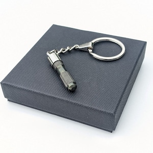 Puede incluir: Un llavero gris metálico con un diseño cilíndrico, con un cuerpo texturizado y un pequeño anillo para llaves. El llavero se presenta sobre una caja texturizada gris oscuro, sobre un fondo blanco.