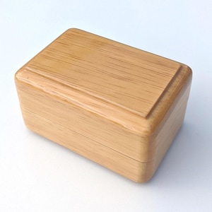May include: A small, rectangular wooden box with a light brown finish. The box has a smooth, polished surface and a slightly raised edge around the top.