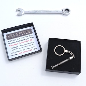 May include: A silver ratchet spanner and a Lotus F1 Team keyring in a black box. The keyring is a miniature actuator stud with a chain and ring. The box contains text about the keyring's origin and the Lotus GP E21 chassis.