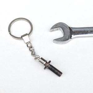 Puede incluir: Un llavero de metal plateado con un perno y detalle de cadena, junto a una llave inglesa cromada. El llavero presenta un pequeño perno con una tuerca y una cadena que se conecta a un llavero. La llave es de tamaño estándar.