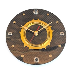 Op de afbeelding: Ronde wandklok met een koolstofvezel wijzerplaat en een goudkleurig tandwielvormig midden. De klok heeft gouden wijzers en een rode secondewijzer. Bevestigd met zilveren schroeven en bouten. Een uniek decoratief stuk.