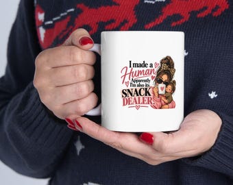Puede incluir: Una taza de cerámica blanca con una ilustración gráfica de una persona con un moño y gafas de sol sosteniendo a un niño pequeño. El texto dice, I made a Human Apparently I'm also its SNACK DEALER, con corazones rosas y texto en rojo.