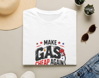 Puede incluir: Una camiseta blanca con un estampado gráfico que incluye el texto "MAKE GAS CHEAP AGAIN" en letras negras y rojas. El diseño presenta una ilustración de una boquilla de surtidor de gasolina roja, una gota amarilla y estrellas pequeñas.