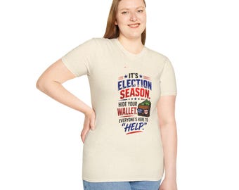 Puede incluir: Una camiseta de manga corta color crema con un gráfico que incluye el texto "IT'S ELECTION SEASON. HIDE YOUR WALLET. EVERYONE'S HERE TO "HELP."" y una ilustración de una cartera marrón con dinero en efectivo y gafas de sol.