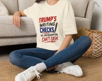 Puede incluir: Una camiseta de manga corta color crema con texto en rojo, negro y azul que dice: TRUMP'S WRITING CHECKS HIS JOB DESCRIPTION CAN'T CASH. El diseño incluye un gráfico de una pluma estilográfica negra junto a la palabra CHECKS.