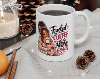 Puede incluir: Una taza de cerámica blanca con una ilustración gráfica de una persona con un moño alto sosteniendo una taza pequeña, rodeada de libros apilados y flores. El texto dice Fueled by COFFEE and MOM GOALS en letras negras y rosas.
