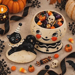 Op de afbeelding: Een keramische Halloween snoepschaal met een lachend maangezicht en een deksel in de vorm van een zwarte kat. De schaal is gevuld met diverse snoepjes, waaronder schedelvormige lekkernijen, oranje pompoenen en kleurrijk verpakte snoepjes.