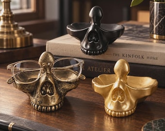 Peut inclure: Trois porte-lunettes en forme de crâne avec des finitions noire, dorée et bronze vieilli. Chaque support présente un pont nasal central conçu pour maintenir la monture d'une paire de lunettes, avec une petite base en forme de plateau.