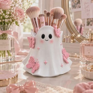 May include: A white ceramic ghost-shaped makeup brush holder featuring raised pink butterfly accents and small star details. The ghost has two black oval eyes and a small smile with pink cheeks, filled with various makeup brushes with pink handles.