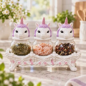 May include: A set of three clear glass spice jars with white and purple unicorn-shaped lids, each featuring a single horn and ears. The jars are held in a white, ornate, rectangular decorative stand with raised floral details and four feet.