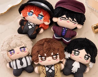 May include: Five small plush dolls with embroidered facial features and distinct hair colours. The dolls wear varied outfits including hats, jackets, ties, and vests in shades of black, brown, tan, and purple, featuring detailed fabric textures.