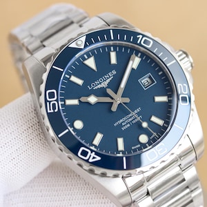 May include: A stainless steel Longines HydroConquest automatic watch with a blue dial and a matching blue rotating bezel. The watch face features luminous hour markers, silver hands, a date window at the 3 o'clock position, and text reading 300M/1000FT.