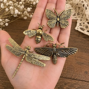 May include: A collection of four antique brass-toned metal insect ornaments featuring a butterfly, a bee, a dragonfly, and a moth. Each piece displays intricate embossed wing details and a textured finish, showcasing realistic anatomical insect shapes.