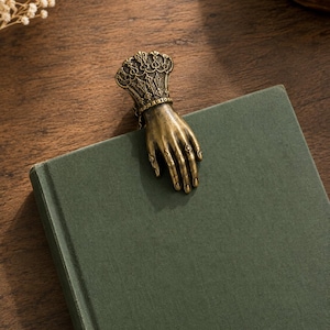 May include: A brass-toned metal bookmark shaped like a hand with an ornate, decorative cuff. The hand is positioned on the top edge of a solid olive green book cover, featuring detailed finger joints and a textured, intricate wrist design.