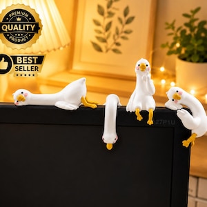 May include: A set of four white goose figurines with orange beaks and feet, each in a different pose. One goose lies flat, one hangs downward, one sits upright, and one leans over the edge of a black monitor frame, all featuring pink cheeks.