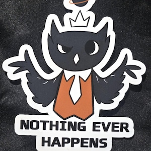May include: A die-cut sticker featuring a black owl character wearing a crown, an orange vest, and a white tie. Above the owl is a small planet with a ring. Below the character, the text reads "NOTHING EVER HAPPENS" in bold black letters.