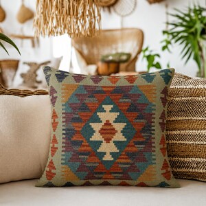 May include: A square throw pillow featuring a woven geometric pattern in shades of sage green, burnt orange, deep blue, and cream. The central design consists of a large diamond shape composed of stacked triangles and stepped motifs.