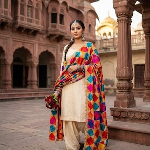 Könnte beinhalten: Ein cremefarbenes Salwar-Kameez-Set bestehend aus einer langärmeligen Tunika und passender Hose, kombiniert mit einem leuchtend bunten Dupatta mit geometrischem Rautenmuster in Rot, Blau, Gelb und Pink sowie einer goldenen Bordüre.