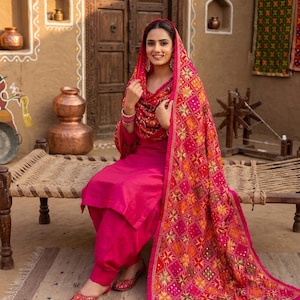 Könnte beinhalten: Ein leuchtend fuchsiafarbenes Salwar-Kameez-Set, bestehend aus einer langärmeligen Tunika und einer passenden Hose. Dazu gehört ein großer, rechteckiger Dupatta mit aufwendigen, bunten geometrischen Stickereien und kleinen reflektierenden Spiegelakzenten.