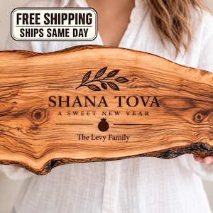 May include: A live edge wooden serving board featuring a dark brown laser-engraved design. The text reads SHANA TOVA A SWEET NEW YEAR and The Levy Family, accompanied by an olive branch illustration and a pomegranate icon centered on the wood grain.