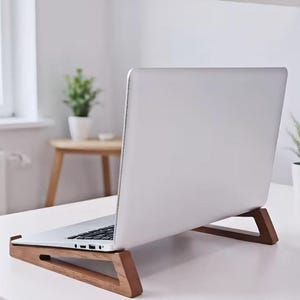 May include: A silver laptop resting on a wooden stand. The stand features a minimalist, angled design with two separate pieces supporting the base of the laptop, elevating it from the white surface to provide an ergonomic viewing angle.