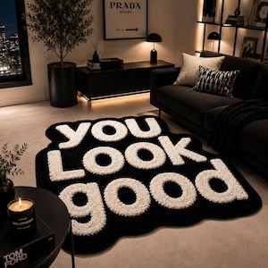以下が含まれることがあります： 波打つような不規則な形の黒いラグで、YOU LOOK GOODという文字が大きく盛り上がった白い質感のある文字で描かれています。ラグは明るい色の床の上に置かれており、部屋には暗い色の家具と温かみのある照明があります。