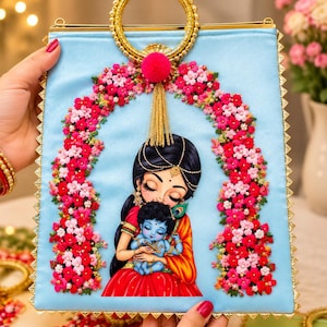 May include: A light blue satin handbag featuring a central illustration of a woman holding a blue-skinned child. The bag is decorated with an arch of pink and red fabric flowers, a gold circular handle, a pink pom-pom, and a gold tassel.
