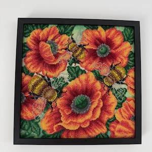 Pode incluir: Uma peça de arte em missangas emoldurada de forma quadrada com papoilas vibrantes cor de laranja e vermelhas com folhas verdes e duas abelhas douradas com corpos texturizados. A obra está numa moldura preta simples com detalhes em missangas.