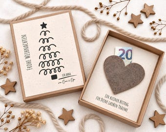 May include: A rectangular cardboard gift box with a lid featuring a black line-art Christmas tree and the text Frohe Weihnachten, Für Anna, and von Oma & Opa. Inside, a grey felt heart sits above the text Ein kleiner Beitrag für deine großen Träume.