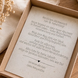 May include: A rectangular cardboard gift box containing a cream-coloured card with black printed German text celebrating an 18th birthday. The card features a heartfelt message, a small heart icon, and the names Carina, Henry, and Dirk at the bottom.