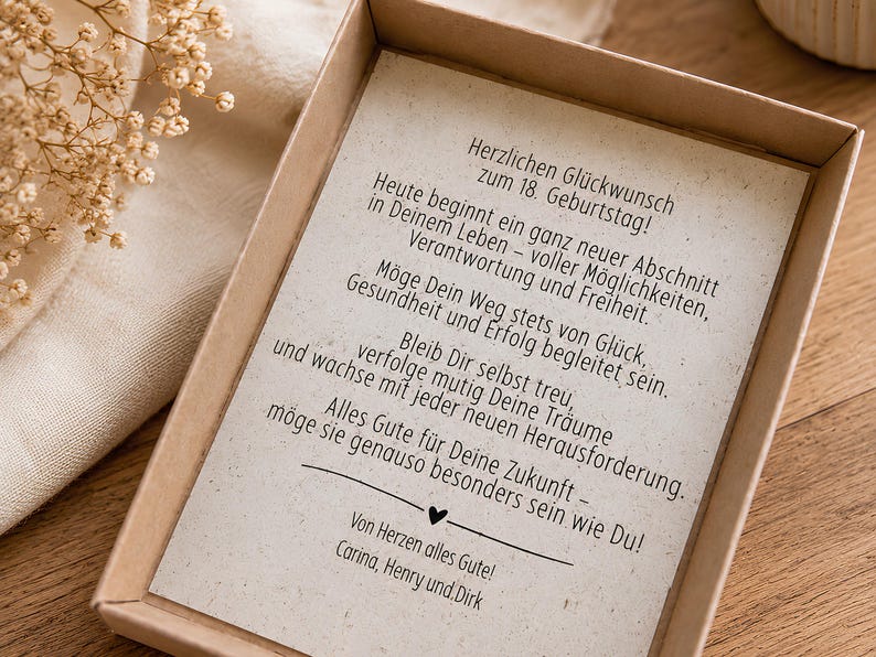 May include: A rectangular cardboard gift box containing a cream-coloured card with black printed German text celebrating an 18th birthday. The card features a heartfelt message, a small heart icon, and the names Carina, Henry, and Dirk at the bottom.