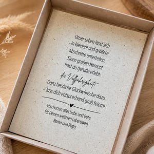 May include: A rectangular cardboard gift box containing a cream-coloured card with black printed German text. The text reads: Unser Leben l&auml;sst sich in kleinere und gr&ouml;&szlig;ere Abschnitte unterteilen. Einen gro&szlig;en Moment hast du gerade erlebt: die Vollj&auml;hrigkeit.